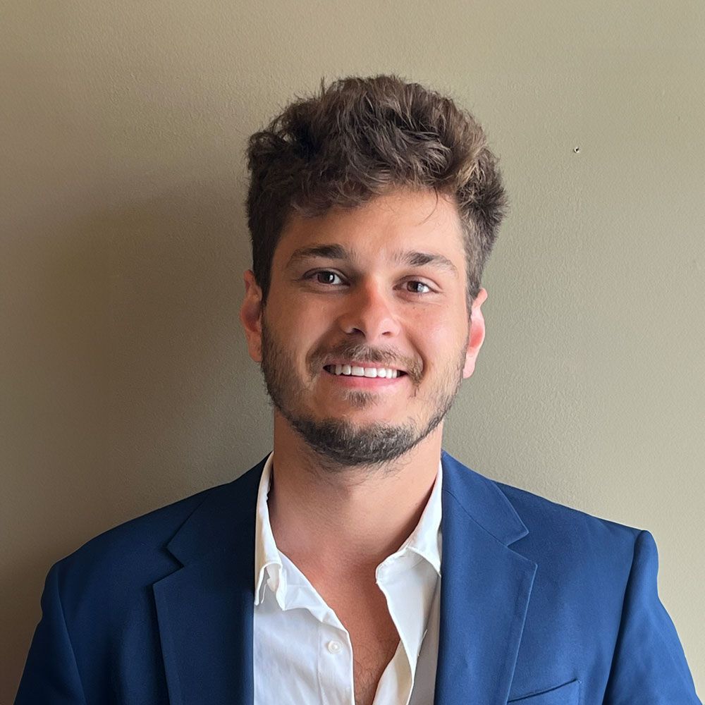 Dr. Khan smiling in a blue blazer and white shirt standing against a plain beige wall.