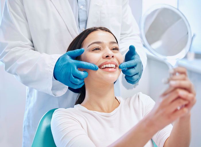 Smiling woman holding a mirror while a dentist in gloves examines her teeth during a checkup.