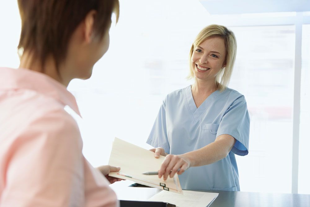 Smiling receptionist in scrubs handing a clipboard to a patient at a front desk in a medical office.