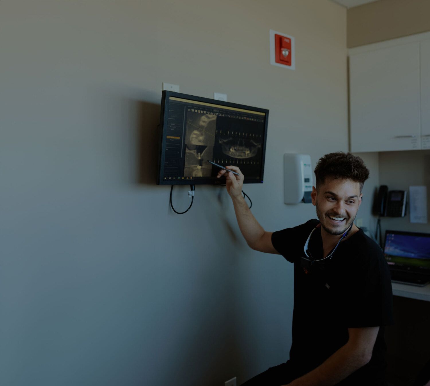 Dr. Khan smiling dentist pointing at a dental X-ray displayed on a wall-mounted monitor inside a modern dental clinic.